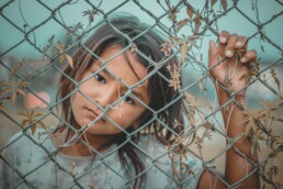 Sad black-haired girl stands behind an overgrown fence.