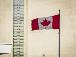 A Canadian flag waves in front of a beige concrete building wall.