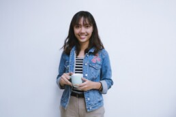 Woman with brown hair wearing a jean jacket holds a cup of coffee smiles